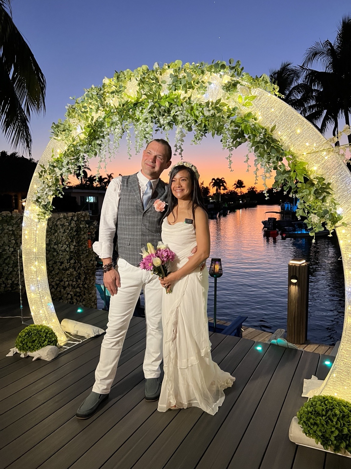 Newlyweds beneath a glowing floral arch beside the waterfront at sunset